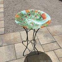 Full view of peacock glass bird bath on metal stand outdoors on stone and gravel