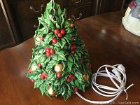 Front and side view of the green ceramic Christmas tree decorated with red berries and gold bells, with electrical cord visible on a wooden surface.