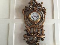 Full view of the large ornate wall clock hanging on a white paneled wall, showing the entire design and frame.