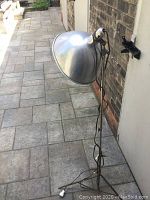 Full view of the vintage adjustable metal utility floor lamp standing outdoors on a paved surface, showing tripod base and shade.