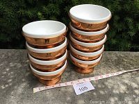Stacked nine copper bowls with white porcelain inserts displayed outdoors on stone surface with measuring tape.