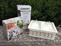 Lot items arranged outdoors with greenery background: boxed Cuisinart soda maker, boxed food mill, and white divided serving dish.