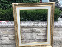 Full view of the large gold ornate picture frame with white matting edge, hung on a stone wall, showing size and style