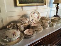 Full view of the collection on a marble surface, showing stacked plates, cups, serving bowls with lids, and various sized pieces with floral pattern.