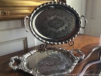 Two silver trays, one held on a stand, showing intricate engraving and handles on both ends.