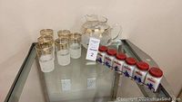 Glass pitcher and six drinking glasses with gold rims displayed on glass shelf alongside vintage spice jars in metal rack.