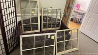 Four vintage wooden windows arranged on and in front of a vintage leather sofa and leaning against a folding screen, showing different pane configurations and white painted wooden frames.