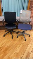 Photo showing both chairs side by side on a wood floor, one black vinyl with armrests and one blue cushion with gray mesh back with no armrests.