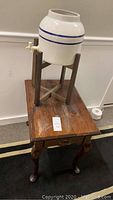 Wooden side table with ceramic jug on stand positioned on top, showing the top and some scratches on the table surface.