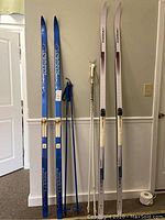 Full view of two pairs of cross country skis and two pairs of poles standing against a wall.