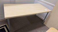 Photo of rectangular table showing light wood finish top surface and silver metal legs against carpeted floor and white wall.