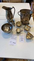 View showing entire lot on white table: metal pitcher, ice bucket, large silver plate bowl, sterling silver baby cup, several small bowls/dishes.