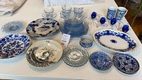 Photo showing the assortment of blue and white china plates, clear glass dish, blue depression glass plates and cups, and two floral print mugs
