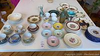 Wide shot of assorted dishware items including tea cups, dessert plates, Italian plates, and vases arranged on a white table.