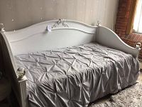 White wooden daybed with curved headboard and footboard, showing full side view with mattress covered in a gray fitted sheet.