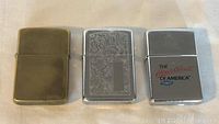 Front view of three Zippo lighters side by side on fabric background, showing design details.