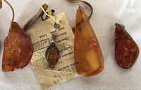 Four amber pendants displayed on white fabric. Includes elongated amber drop pendant, dark amber teardrop pendant, irregular amber pieces with leather cords, and a leaf-shaped amber pendant set in a silver frame.