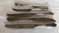 Top view of two cheese knives and three dinner knives with ornate sterling silver handles and stainless steel blades
