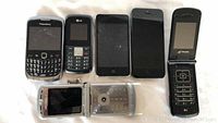 Overview of all cell phones and iPods in lot on white fabric, showing a range of BlackBerry, LG, iPhone, Motorola, Telus devices and iPods, all with varying wear and visible dirt or scratches.