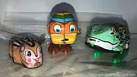 Photo showing front view of three tin toys in plastic bags: a brown mammal, an orange fish/duck-like toy, and a green frog toy.