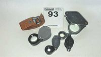 Top view of three metal jeweler's loupe magnifiers and a small brown leather case on white background.