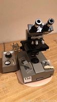 Front view of microscope body on table, showing binocular head, stage, objectives, transformer unit, and eyepieces in plastic bags on base