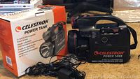 Photo of the Celestron Power Tank in front of its original packaging box. Shows the front panel with controls, switches and the 12V DC output port. The spotlight is visible on the left side of the device. A black power cable is attached.