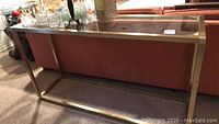 Side angle view showing brass frame and glass top of console table with background furniture.