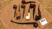 Eight tobacco pipes displayed on beige cloth: includes smooth, rusticated, sandblasted and ceramic bowls plus a long churchwarden.