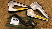 Wide shot showing two white Meerschaum pipes in black cases with yellow stems alongside a black carved pipe resting on a plaid cloth.