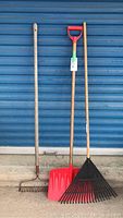 Wide view showing all three tools (metal rake, red plastic shovel, leaf rake) leaning against blue garage door.