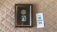 Framed display showing coin and stamp on black background, with visible seller's MaxSold lot number card.