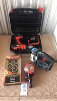 Full lot view showing Black & Decker cordless drill in open carrying case, portable work light, brass floor grate in box, and battery pack on table.