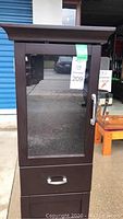 Full front view of the dark brown wooden cabinet showing the glass door and drawer with metal handles.