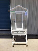 Front view of white metal birdcage standing on wheels outside a building, closed door visible on cage front