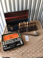 Blue metal tool box, wood-handled chisel set in leather roll, 40-piece ratchet socket set in green box, and large metal crowbar laid out on table.