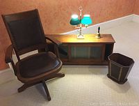 Full view of office set including leather padded wooden swivel chair, oak desktop bookcase with sliding glass doors, brass lamp with two green fluted glass shades on marble base, wood-handled brass bell, and gilt embossed leather wastebasket.