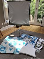 Full view of the Beat Pad, Wii Fit board, cables, manuals, and the projector screen on tripod stand in a room.