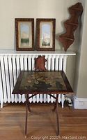 Overview of folding wood table with floral decoupage top, two framed prints positioned behind, and wall-mounted shelf piece