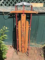 Full view of the folded vintage Flexible Flyer sled leaning against a lattice fence. Shows wooden slats, red metal frame, and rope handle for steering.