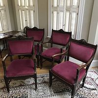Four ornately carved armchairs with lion head and floral motif wooden frames. Upholstered in wine-colored velvet with double piping. Positioned on a floral area rug in front of window shutters.