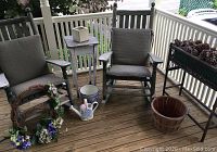 Photo showing two weathered wooden rocking chairs with taupe cushions, small shabby chic table, outdoor wreath, ceramic planters, and rectangular wicker plant stand containing pine cones