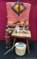 Front view of the woven child's poncho, three statuettes, a small tom tom drum with drumstick, and rustic wooden stool arranged on a red cloth background.