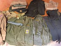 Overview of multiple atomic U.S. Army uniform pieces including fatigues, dress blues jacket, khakis, cap, patches, and buttons displayed on floor
