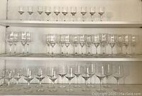 Shelf with three rows of stemware showing a variety of clear glass stemmed glasses including aperitif glasses, wine goblets, and other stemmed glass types in different heights and patterns.