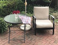 Patio outdoor scene showing folding glass-top table with plaid tablecloth, glass vase with red fabric flowers, and patio chair with beige cushions