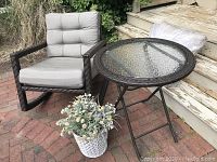 Patio furniture set on brick patio with brown resin rocker, gray cushions, folding glass top side table, and white woven basket with artificial flowers