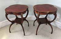 Pair of two clover-shaped leather-top end tables with mahogany veneer trim and gilded embossed borders, bowed legs with gilt accents, and lower shelves. Damage visible on one leather top.