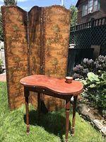 Three-panel folding decorative screen with pastoral scenes and kidney-shaped wooden table with drawer, small brown ceramic pot on table, setting outdoors on grass