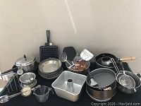 Photo showing assortment of cookware and utensils arranged on floor including pans, cake pans, colander, waffle maker, mixing bowls, and measuring cup